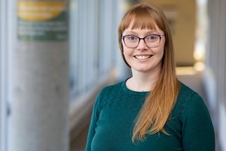 Portrait of Governor Heather Wiebe, smiling, with long red hair, wearing a green shirt and glasses in an airy, modern room. 